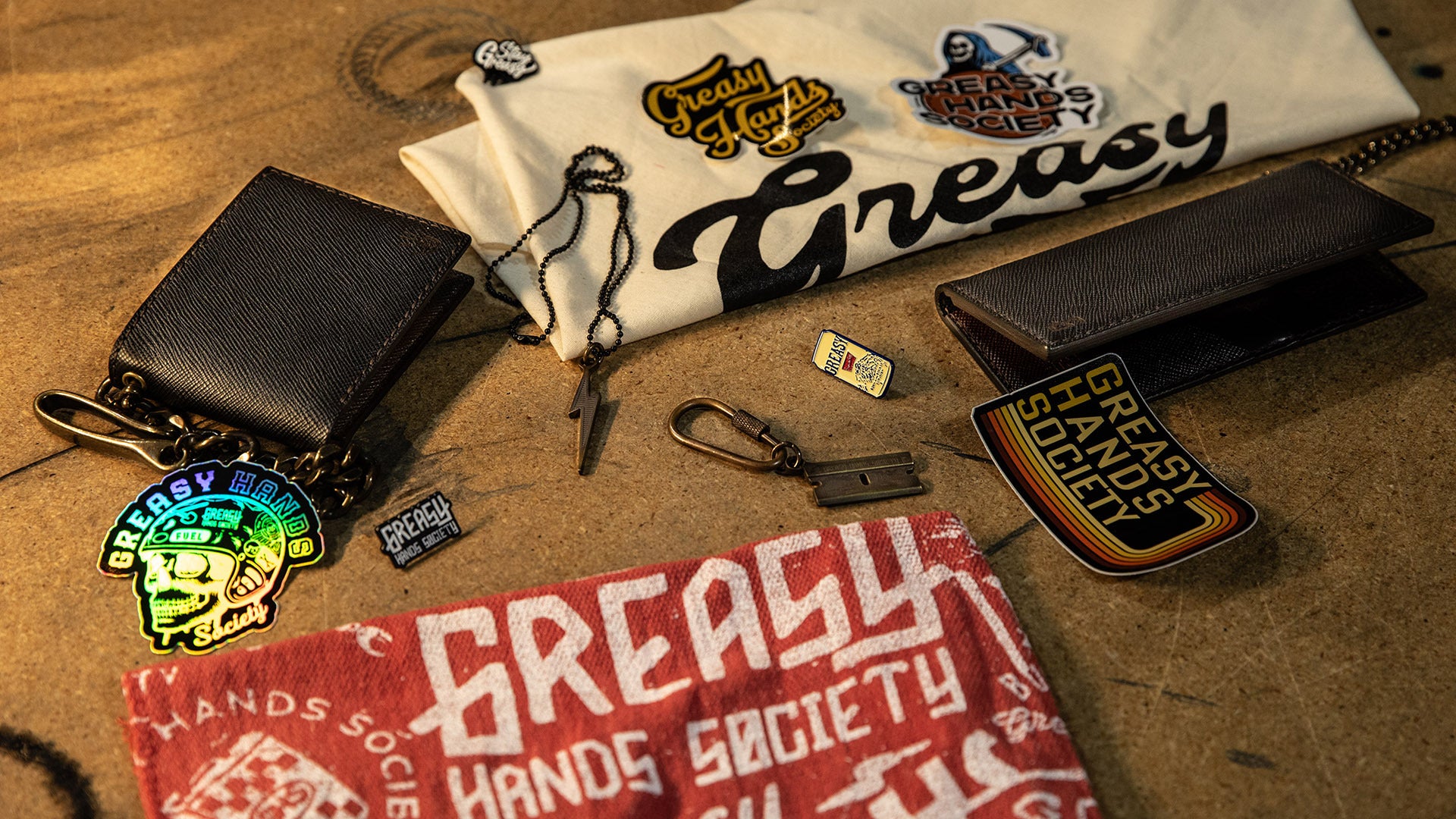Collection of Greasy Hands Society merchandise including a t-shirt, wallet, keychain, and patches on a wooden surface.